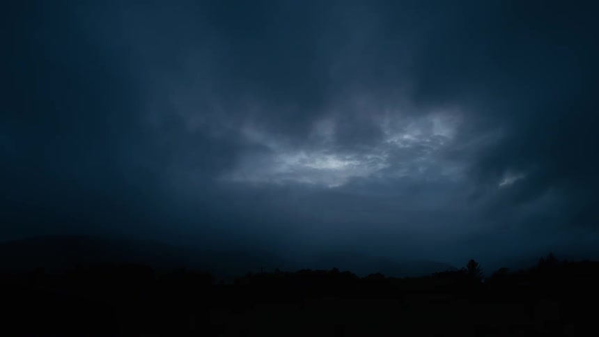 A mysterious, dark blue sky is filled with ominous clouds at twilight, with a faint light breaking through the center