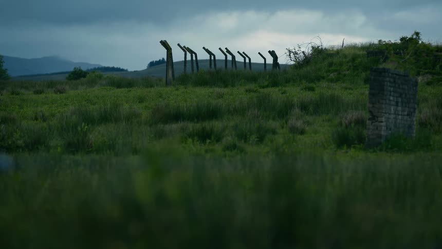 Ominous concrete fence posts and ruins stand on a grassy hill under a dark, stormy sky