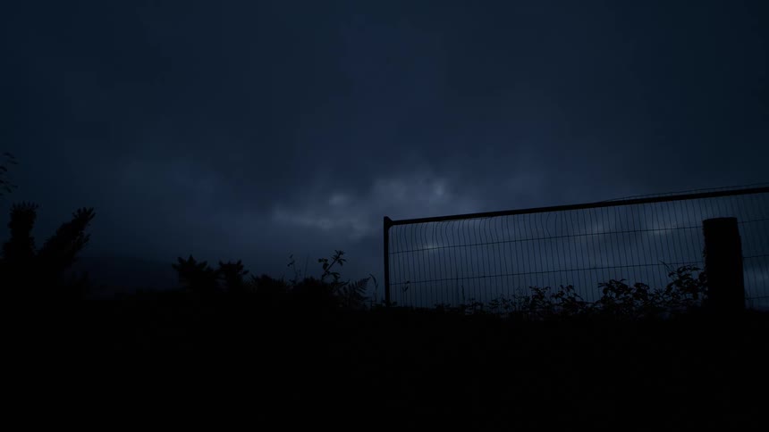 Lightning illuminates a dark, stormy sky above a silhouetted fence at night