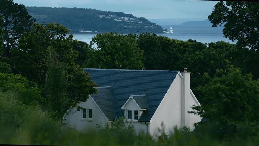 Birds fly over a tranquil coastal home nestled in a dense forest, with a view of the water and a distant lighthouse