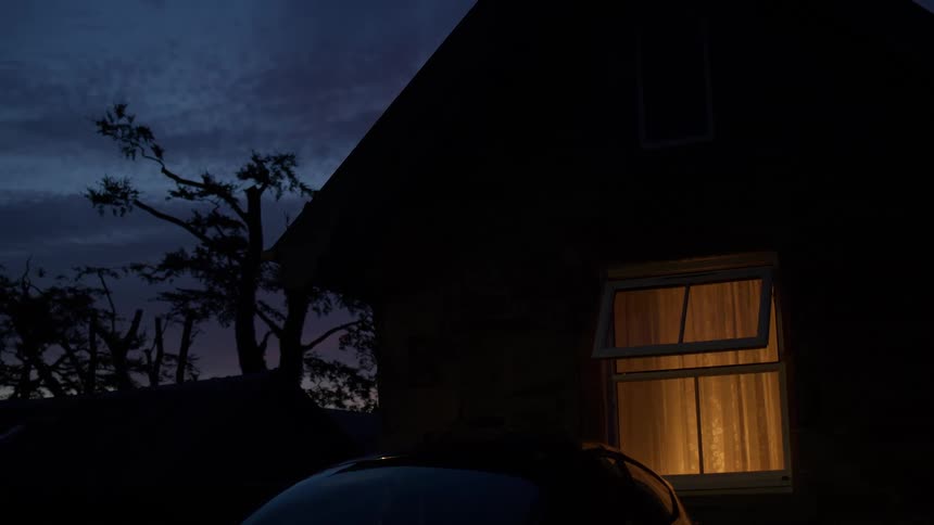 A warm light glows from a single open window of a house at dusk