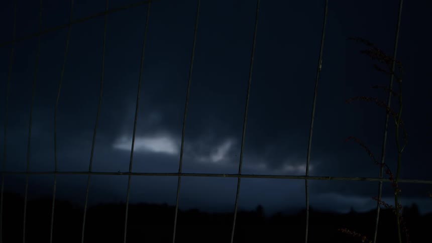 Dark storm clouds loom in the night sky behind a wire fence and a silhouetted plant
