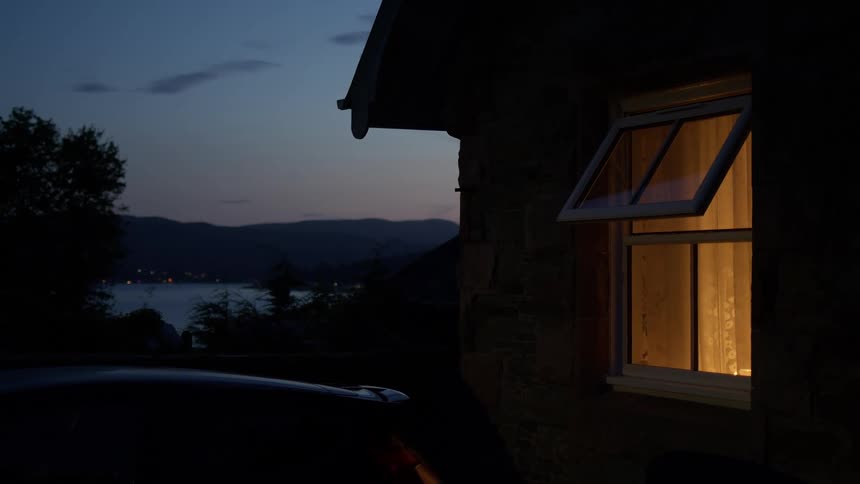 A warm light glows from an open window of a stone cottage at twilight, with a view of a loch and mountains in the distance