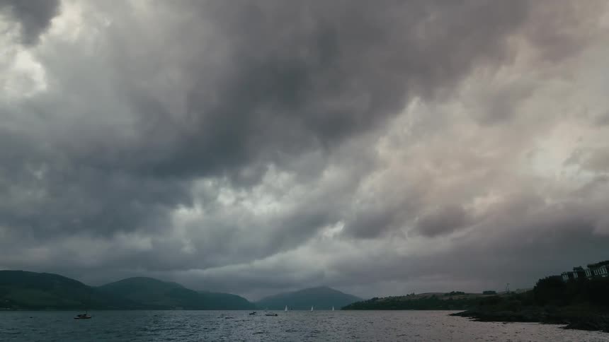 A dramatic timelapse shows stormy clouds moving over a loch with distant mountains as sailboats navigate the choppy waters