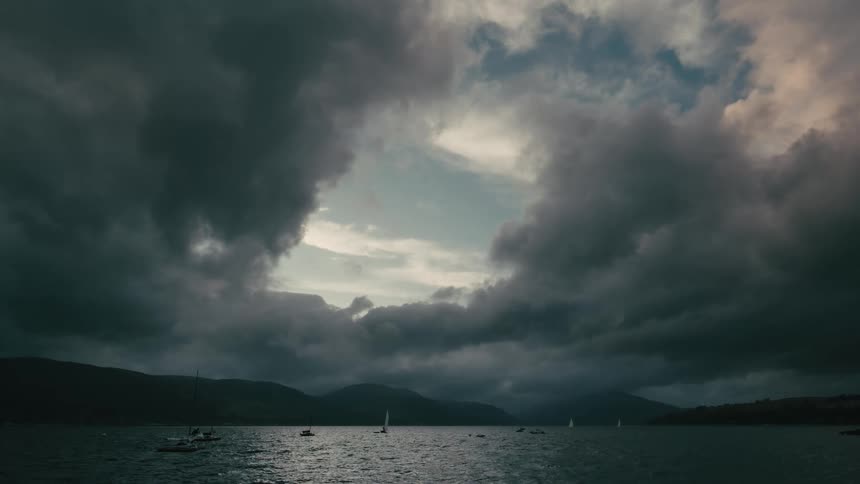 An eye in the storm clouds hangs over sailboats on a vast, dark lake surrounded by mountains
