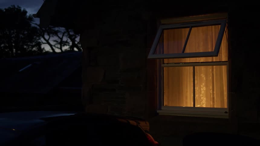 A warm light glows from inside an open window of a stone house at dusk