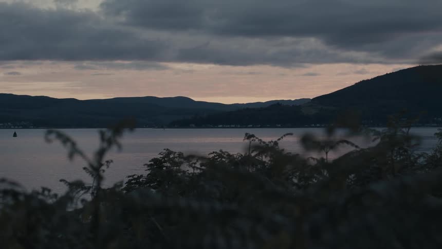 A serene, moody view over a calm lake towards distant mountains under a cloudy sky at dusk