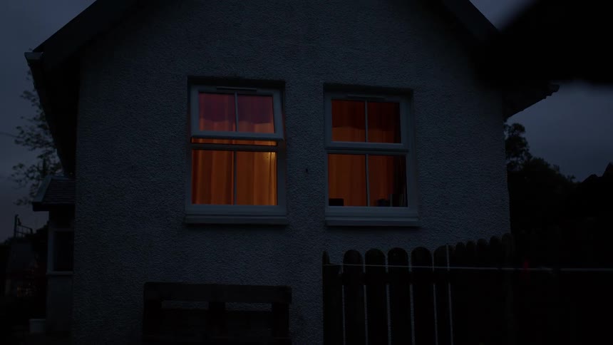 Warm orange light glows from two windows of a small house on a dark, overcast evening