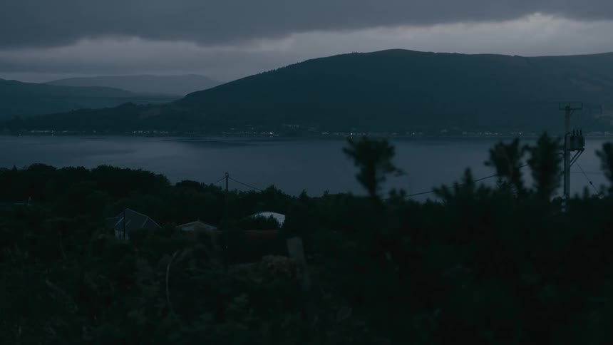 A serene and moody view of a Scottish loch at dusk, with lights from a distant village twinkling across the water