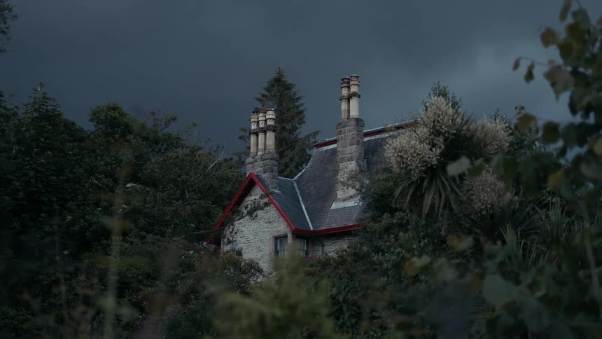 An old stone cottage with red trim is nestled amongst dense trees under a dark and stormy sky