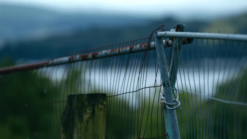 Close up of a rustic metal fence post overlooking a tranquil lake and hills on a moody, overcast evening