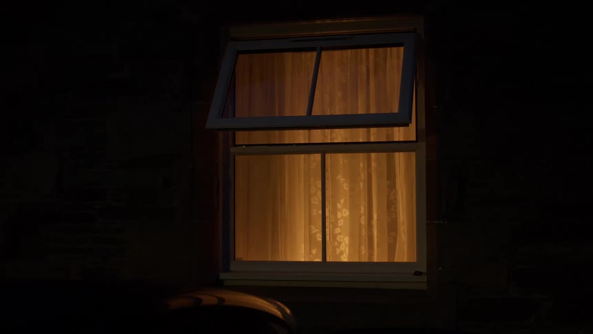 Exterior view of an open window with warm light shining through the curtains at night