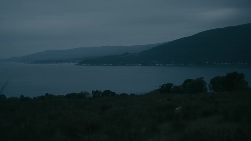 A wide shot of a tranquil loch and distant village with twinkling lights, framed by dark mountains at dusk