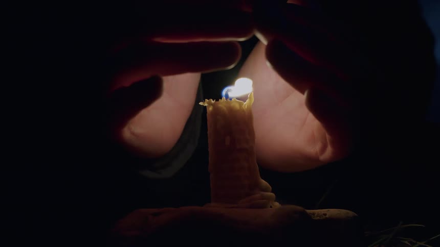 A pair of hands cup around a flickering wax candle, protecting the flame in the complete darkness
