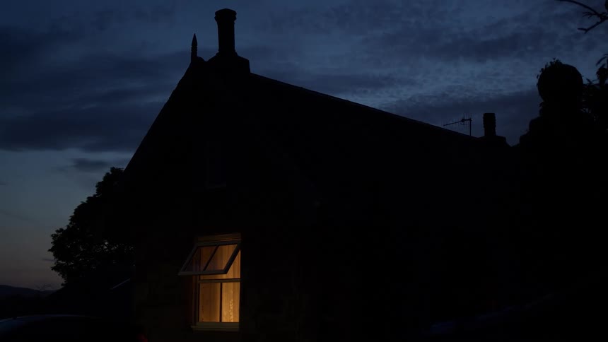 A single warm light glows from an open window of a silhouetted country house at dusk