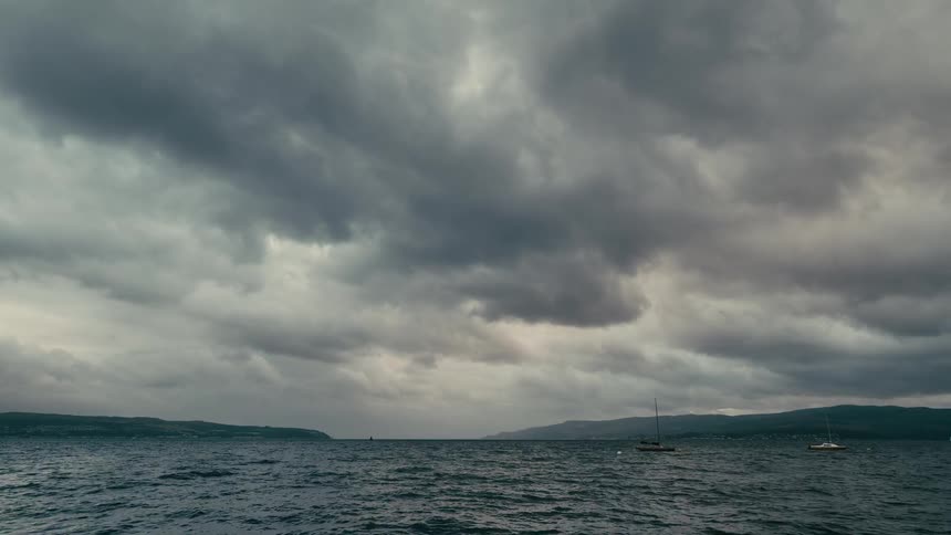 Sailboats are anchored in a bay as a dramatic storm with dark, heavy clouds rolls in over the choppy water