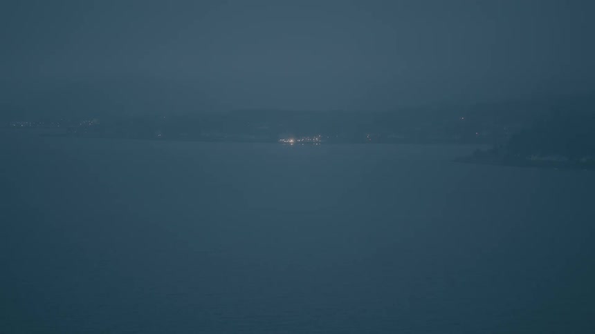 Distant city lights shimmer across a vast body of water on a dark, misty evening