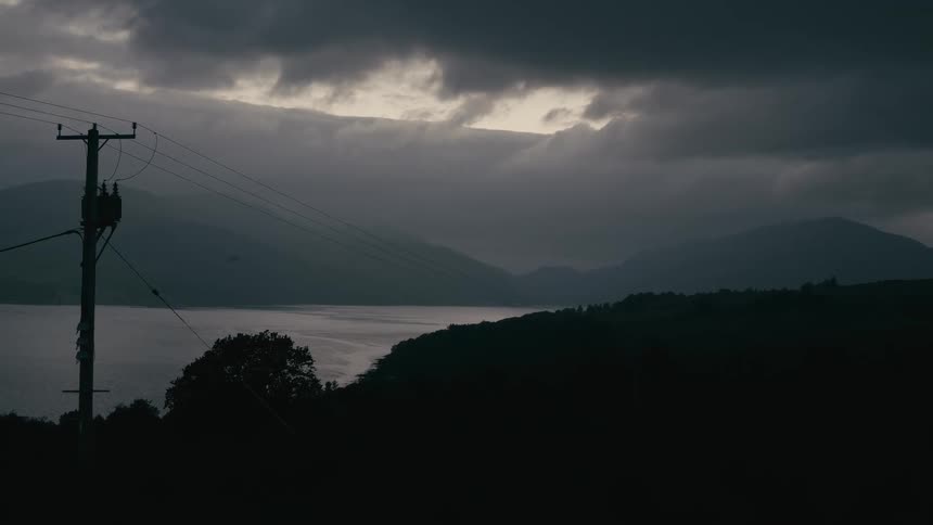 A dark and moody landscape of a lake and mountains under heavy storm clouds at dusk
