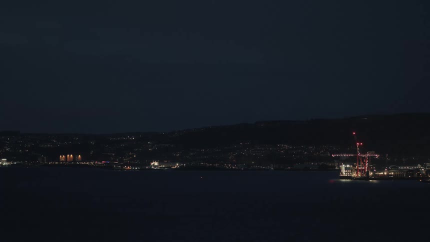 A wide, static shot of a coastal city at night, with its lights twinkling across the water and a large illuminated crane in the harbor
