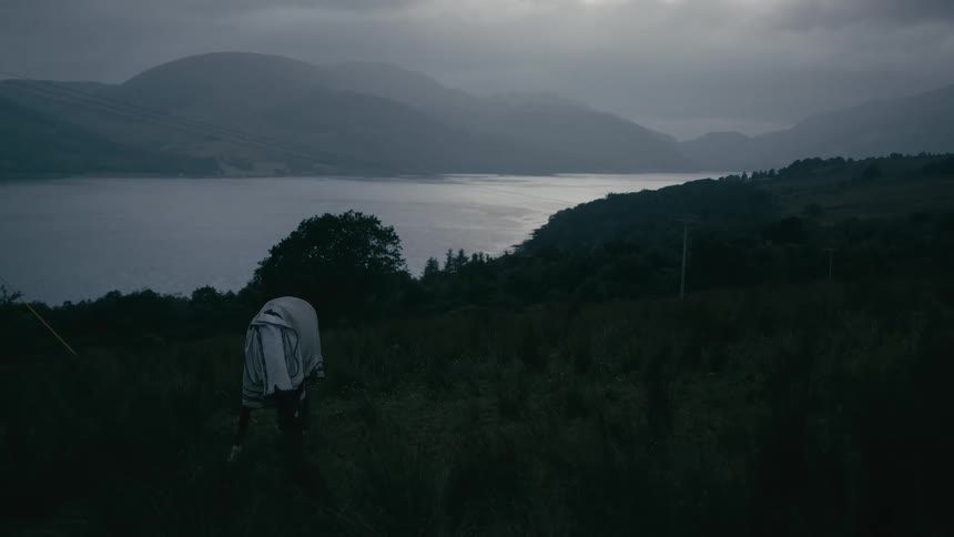 A lone scarecrow stands in a dark, grassy field overlooking a vast lake and mountains on a gloomy, overcast evening