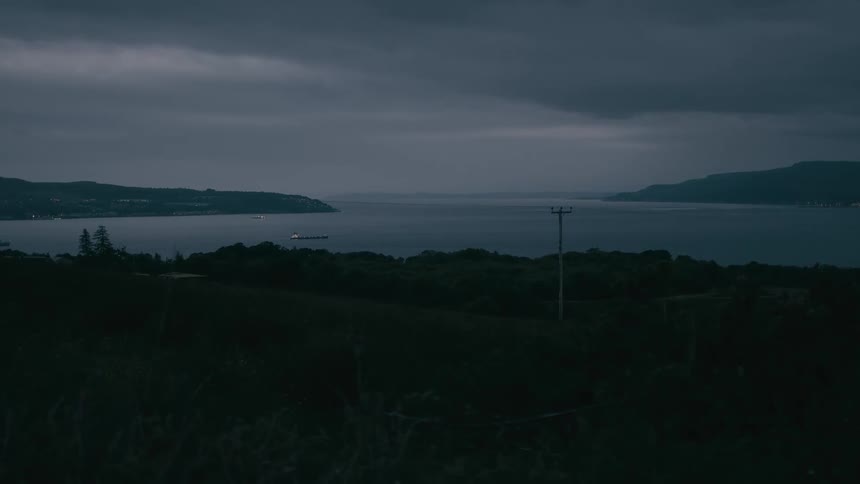 A large ship with its lights on sails across a vast fjord at dusk under a dark and cloudy sky