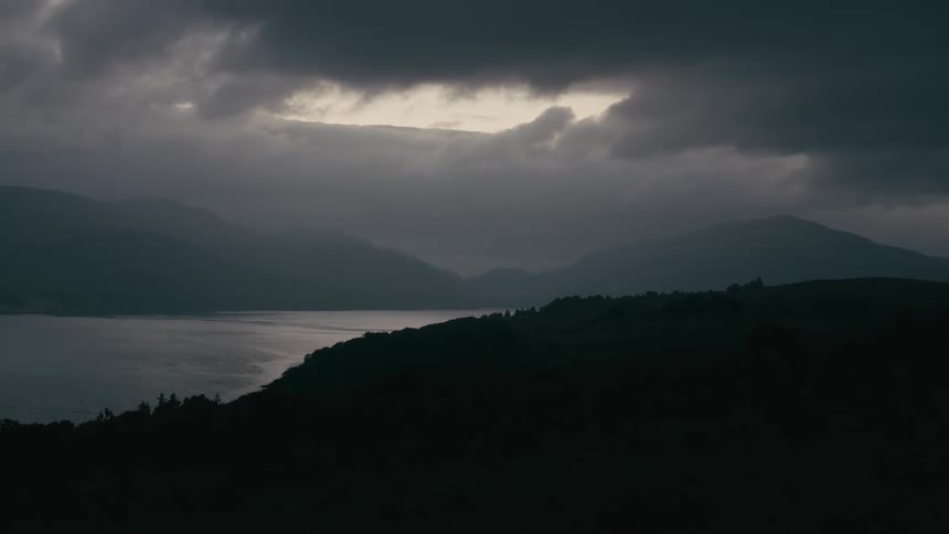 A sliver of light breaks through dark, heavy storm clouds over a tranquil lake surrounded by silhouetted mountains