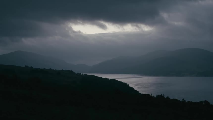 A sliver of light breaks through the dark, stormy clouds over a tranquil loch surrounded by silhouetted mountains