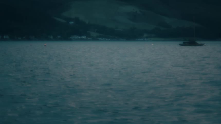 Raindrops fall on a dark, moody lake with a single boat anchored near a tree-covered hillside at dusk