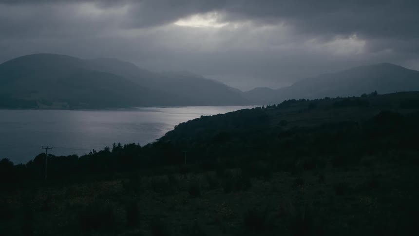 A dramatic and moody landscape shows a lake surrounded by hills under a dark, cloudy sky at dusk