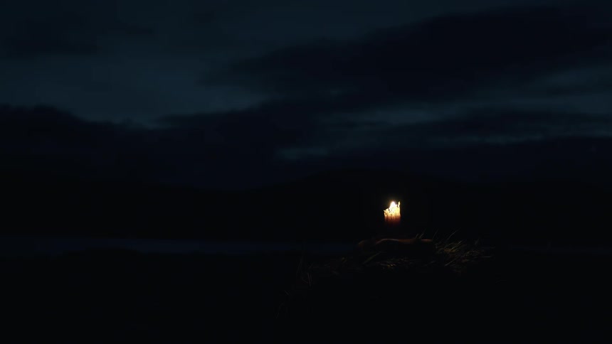 A solitary candle flickers in the wind against a dark and cloudy night sky, its flame the only source of light
