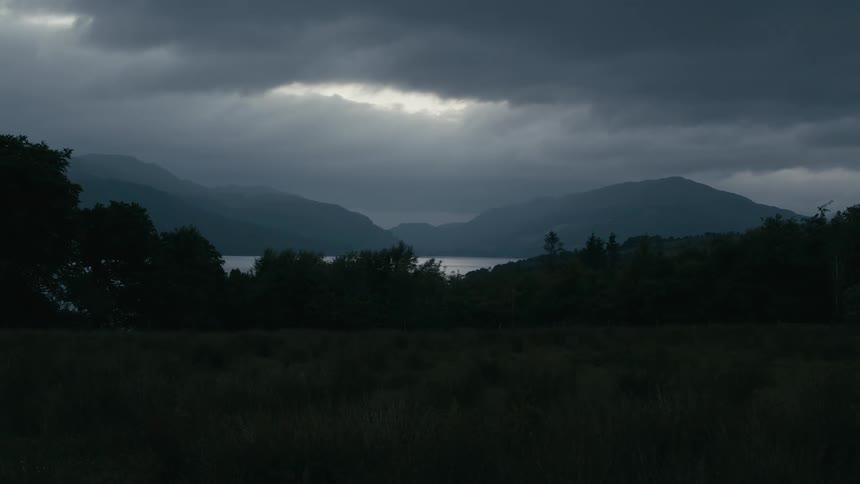 A beam of light breaks through dark storm clouds over a remote lake surrounded by mountains