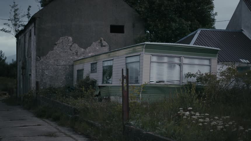A derelict and forgotten mobile home sits next to a crumbling stone building in an overgrown, eerie rural setting