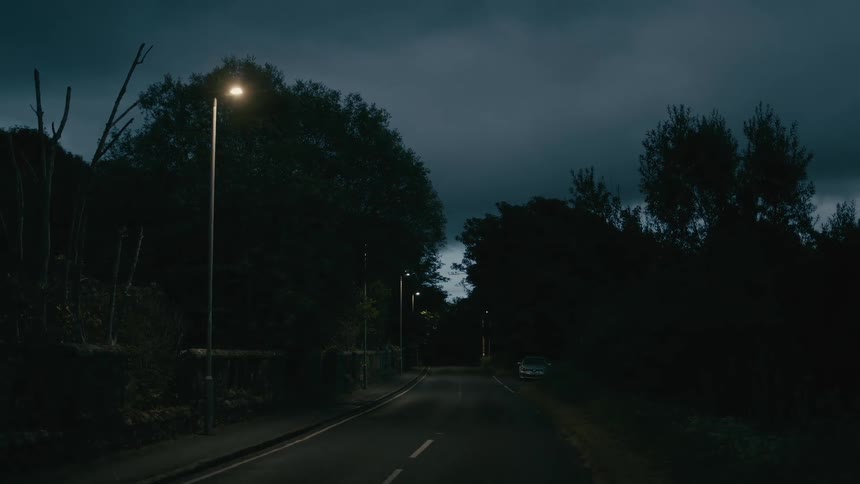 An eerie, deserted road winds through a dark forest at night under a cloudy sky