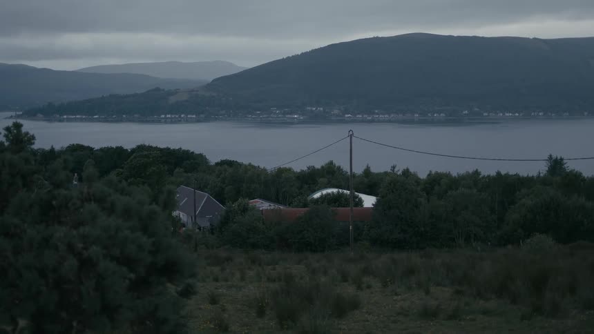 Dusk settles over a tranquil lake and a distant town, with lights beginning to twinkle on under an overcast sky