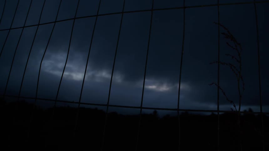 Ominous storm clouds gather in the night sky, viewed from behind a silhouetted wire fence
