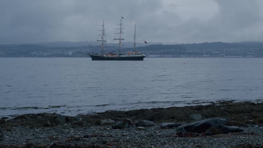 A majestic tall ship is anchored offshore on a dreary, overcast evening, viewed from a rocky beach