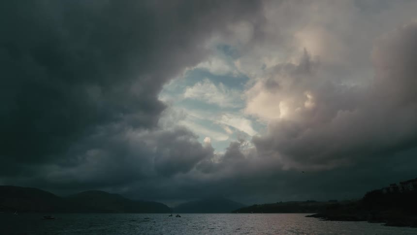 A lone bird slowly flies across an opening of blue sky surrounded by dark, dramatic storm clouds over a calm mountain lake