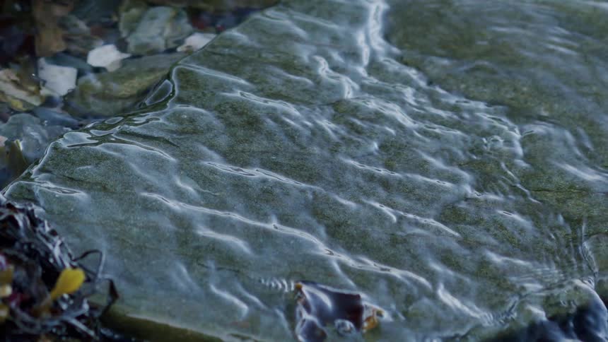 Calm, clear water washes over a smooth rock in slow motion on a pebbled shoreline