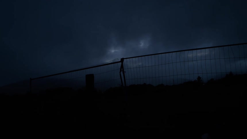 Ominous lightning flashes illuminate dark storm clouds moving across the sky at night behind a silhouetted fence