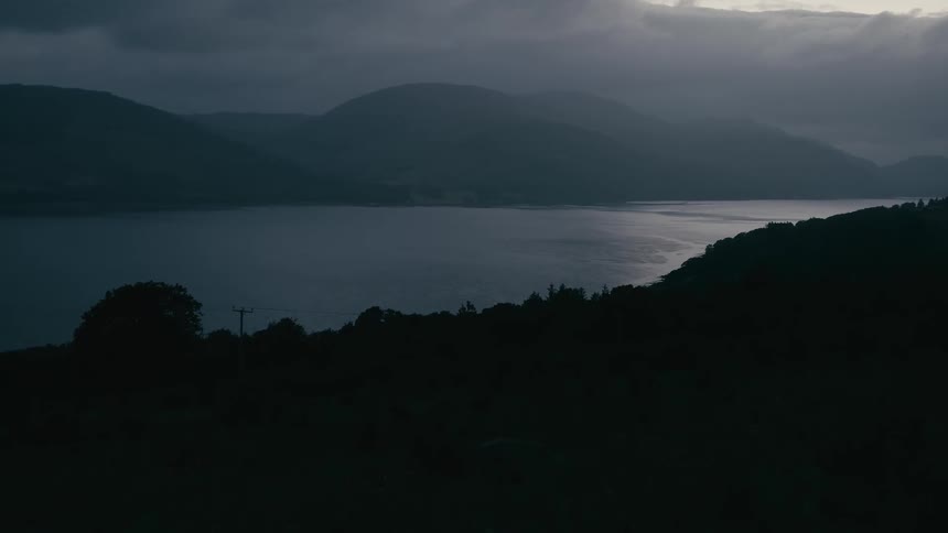 A moody wide shot of a tranquil loch nestled between dark, silhouetted mountains at twilight