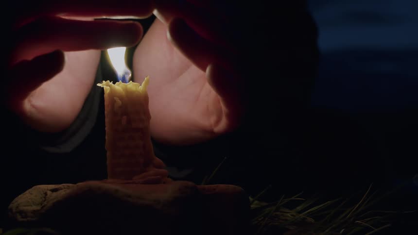 A close-up of hands cupping around a flickering lighter flame to light a textured beeswax candle in the darkness