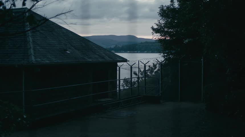 A dark and foreboding shot of a building behind a barbed wire fence, overlooking a body of water at twilight