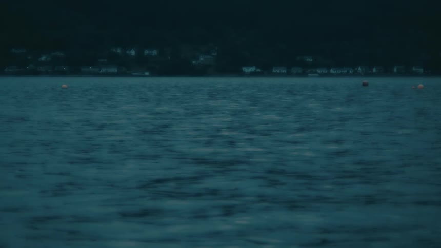 A still shot of calm, dark blue water rippling gently at night with a distant town on the shoreline