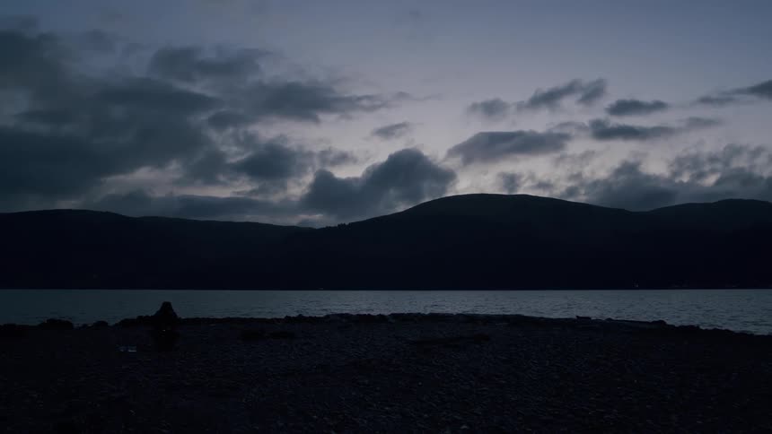 A lone traveler sits by a vast, tranquil loch at dusk, lighting a small lantern on the rocky shore as darkness falls over the mountains