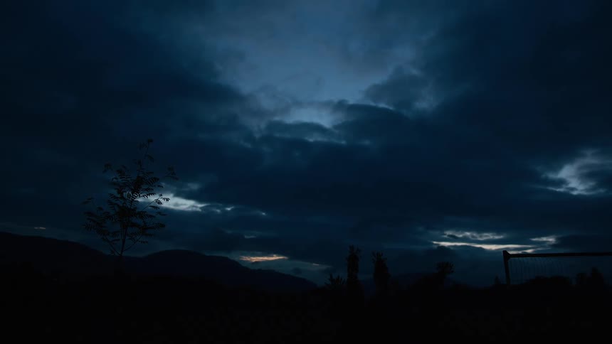 An ominous dark blue sky filled with clouds hangs over a silhouetted mountain landscape just after sunset