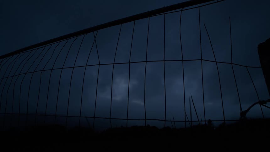A dramatic thunderstorm flashes with lightning behind a silhouetted, broken wire fence at night