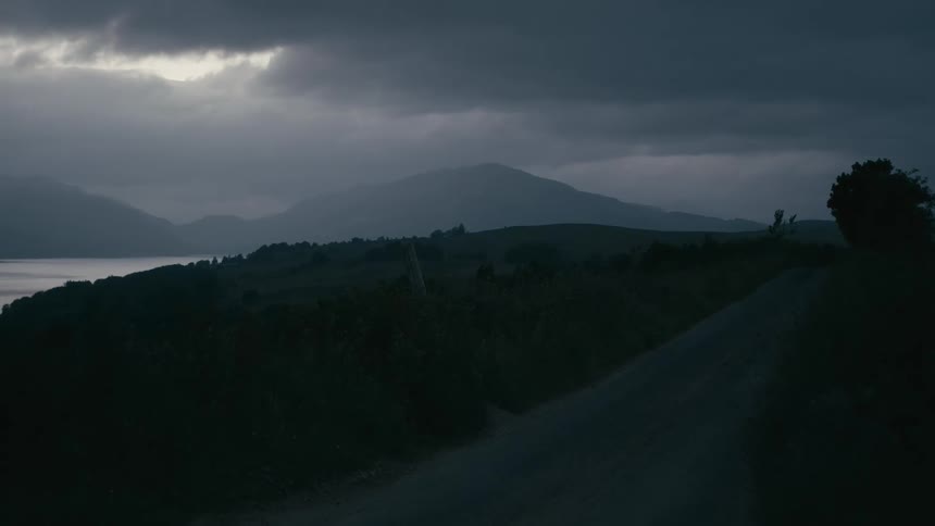 A desolate country road winds through rolling hills next to a lake under a dark, stormy sky at dusk