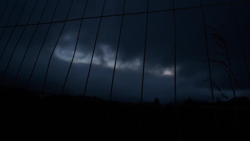 A static shot looks through a metal fence at a dark, moody sky with ominous clouds moving slowly overhead