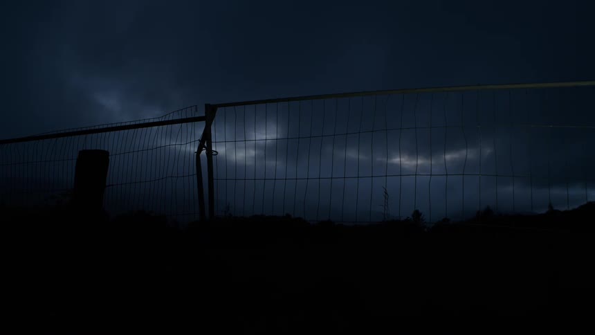 A silhouetted wire fence stands against a dark, stormy night sky with hints of light breaking through the clouds