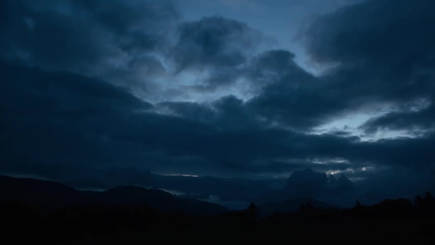 Dark, moody clouds slowly pass over a silhouetted mountain range as evening falls